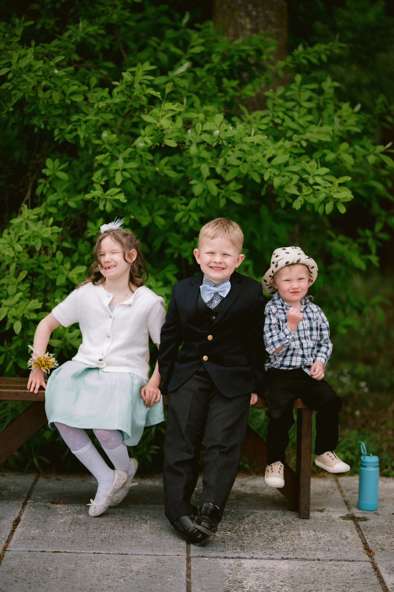 Three children sit and laugh together on a wooden bench in front of green foliage.