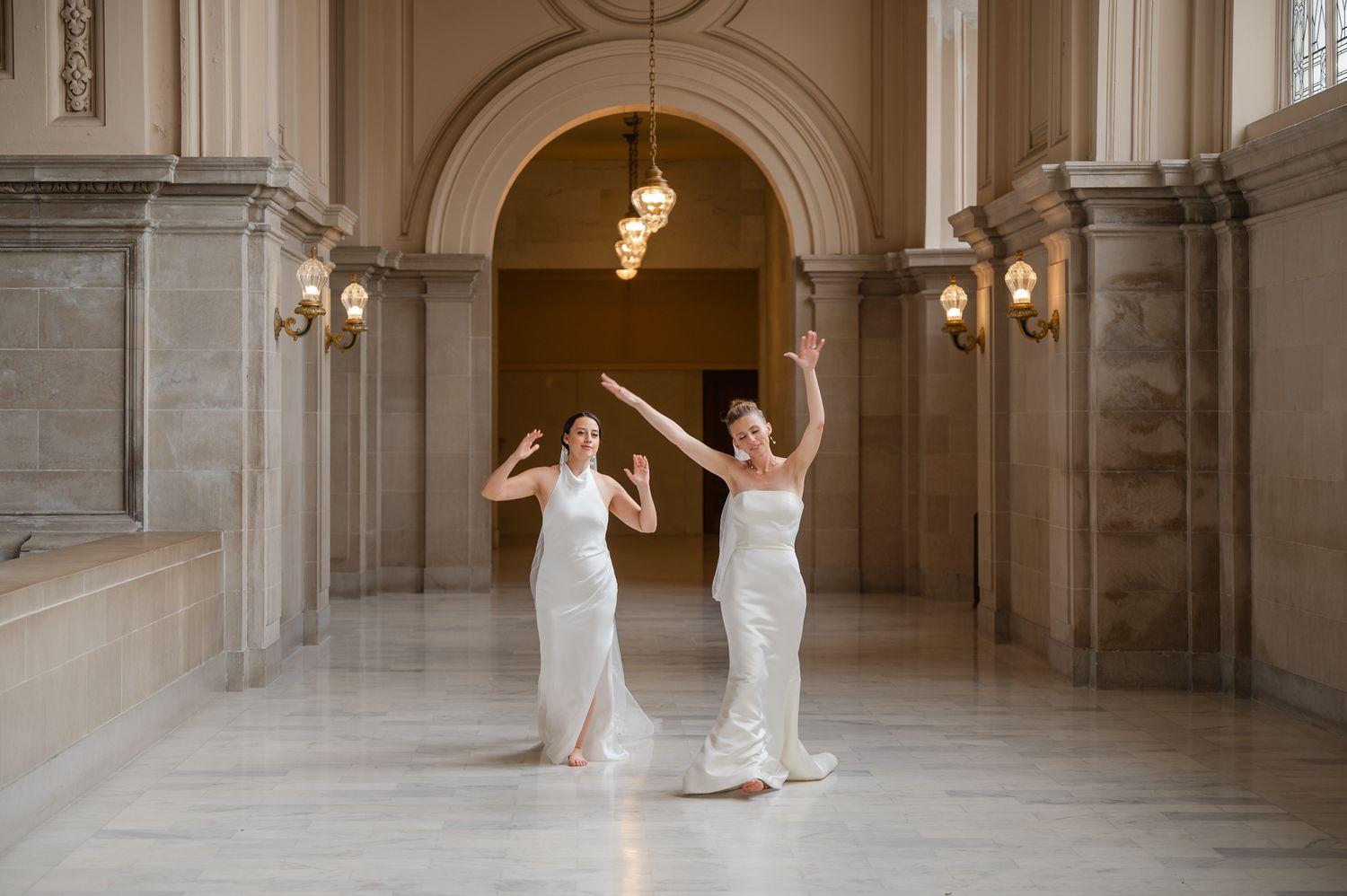 Two people in flowing white dresses dance gracefully in an elegant marble hallway with arches and wall sconces.