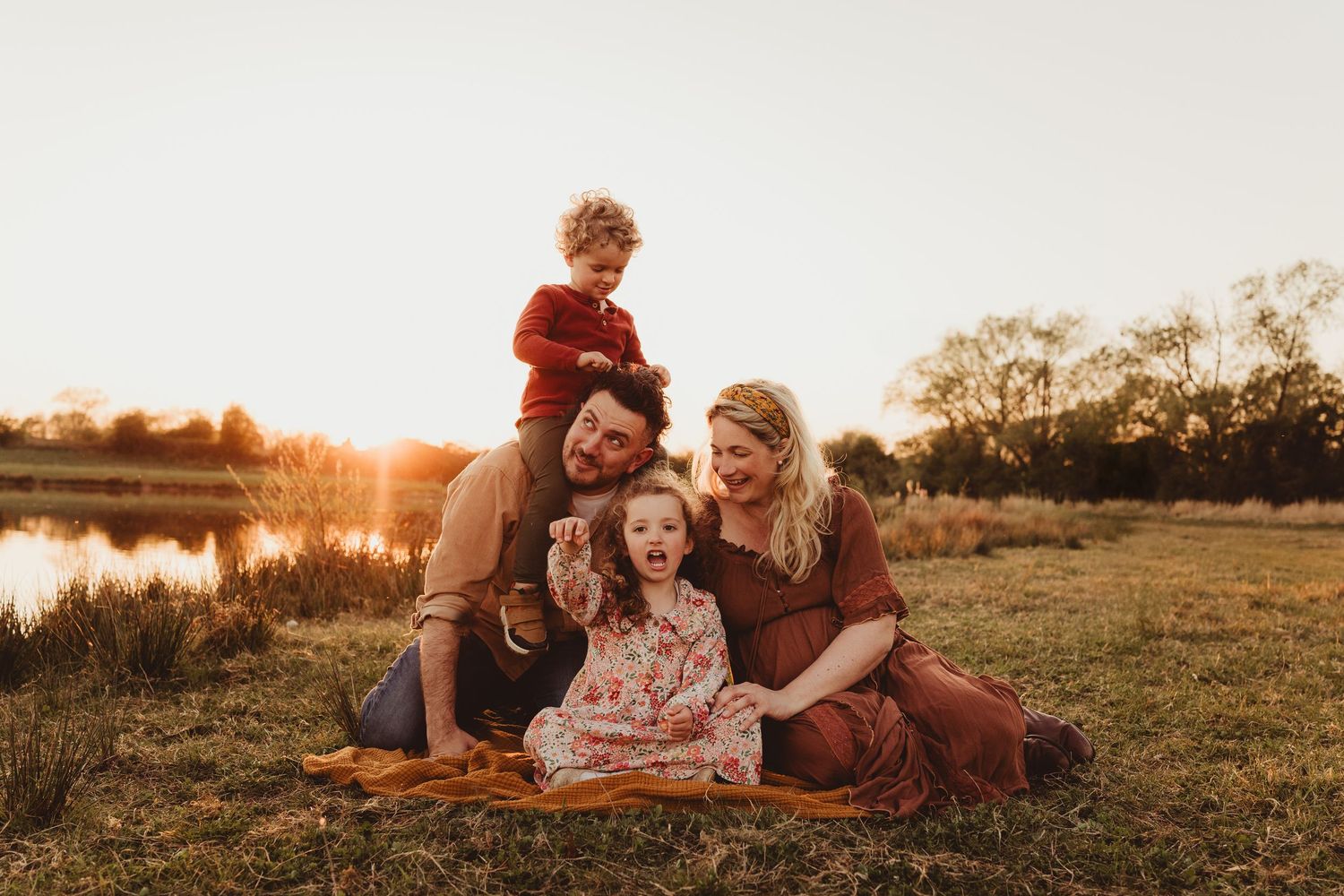 Family enjoys sunset moment together on grassy lakeside bank during golden hour in autumn setting.