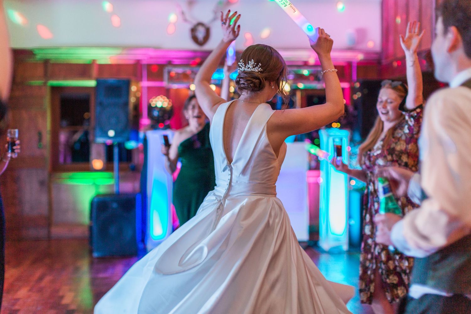 A bride in a white wedding dress twirls on a dance floor illuminated by colourful pink and green party lights at Deer Park Hall Cotswolds