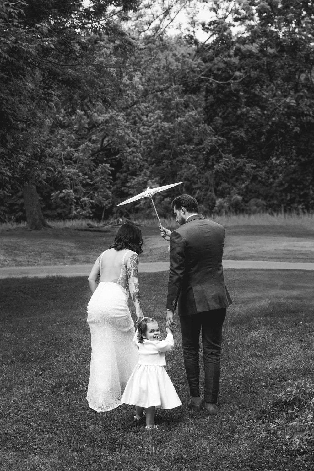 A couple stands with a small child in a white dress under an umbrella on a grassy lawn surrounded by trees.