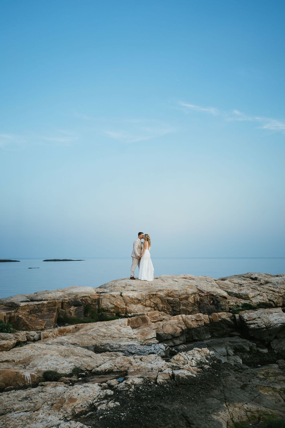 A couple stands together on rocky coastal cliffs overlooking a calm ocean under bright blue skies.
