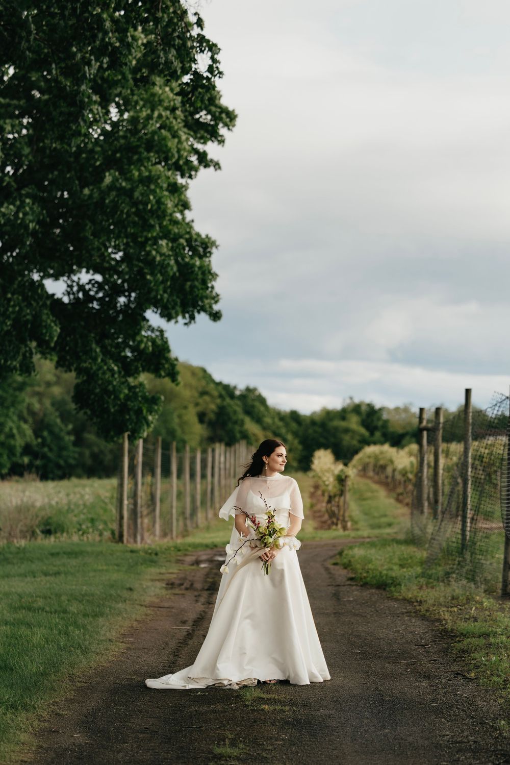 A figure in a white wedding gown holds a bouquet while standing on a dirt path between vineyard rows on a cloudy day.