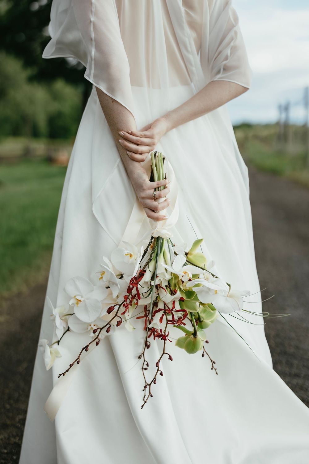 A person in a flowing white dress holds a delicate arrangement of white orchids and greenery against a natural outdoor backdrop.