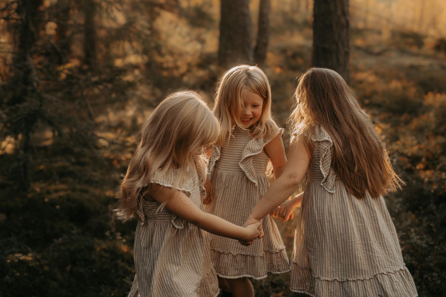 Three young girls in vintage-style linen dresses share a playful moment together in a sunlit autumn forest setting.
