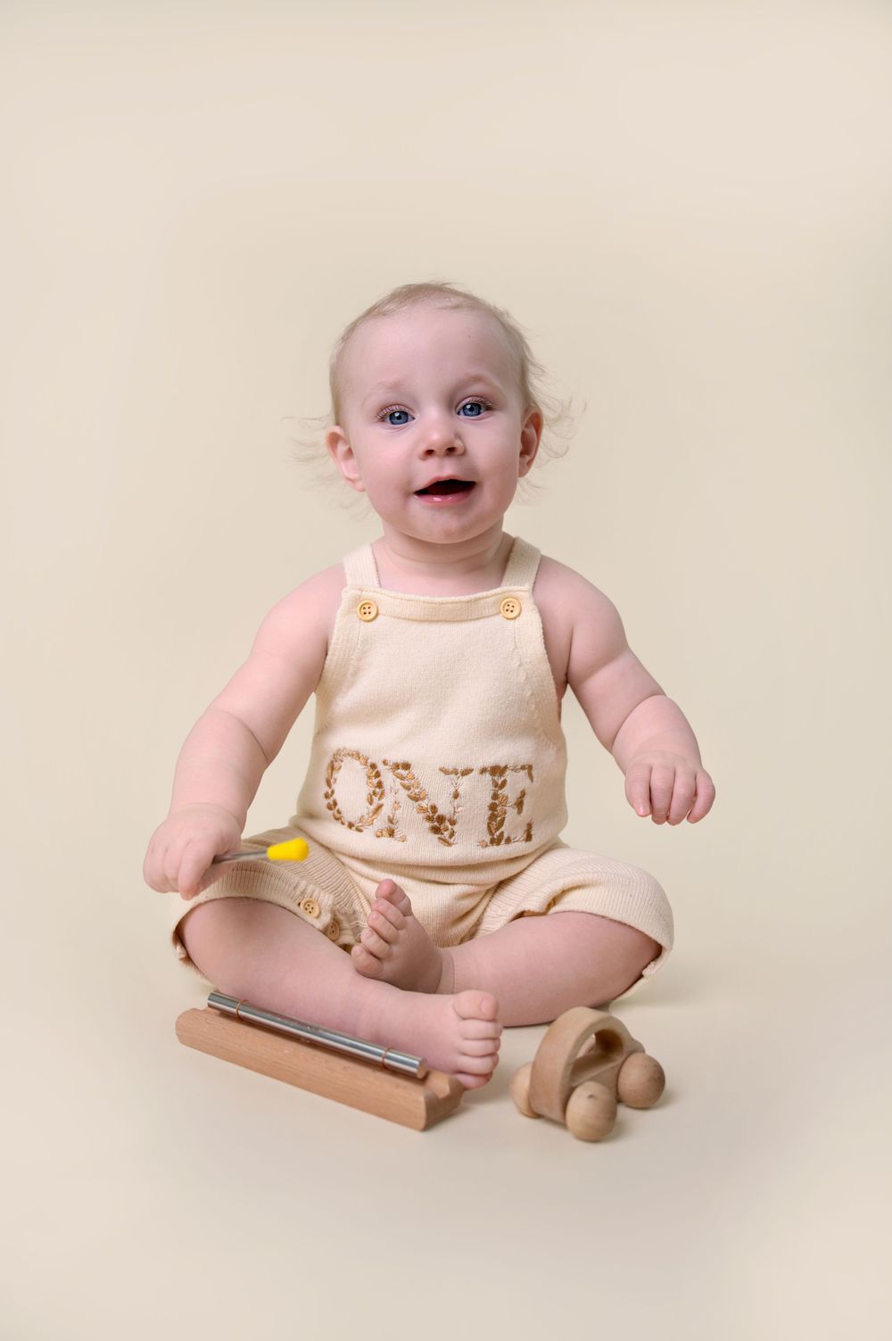 Baby wearing cream-colored romper with "ONE" text sits on wooden surface with a toy beside them.