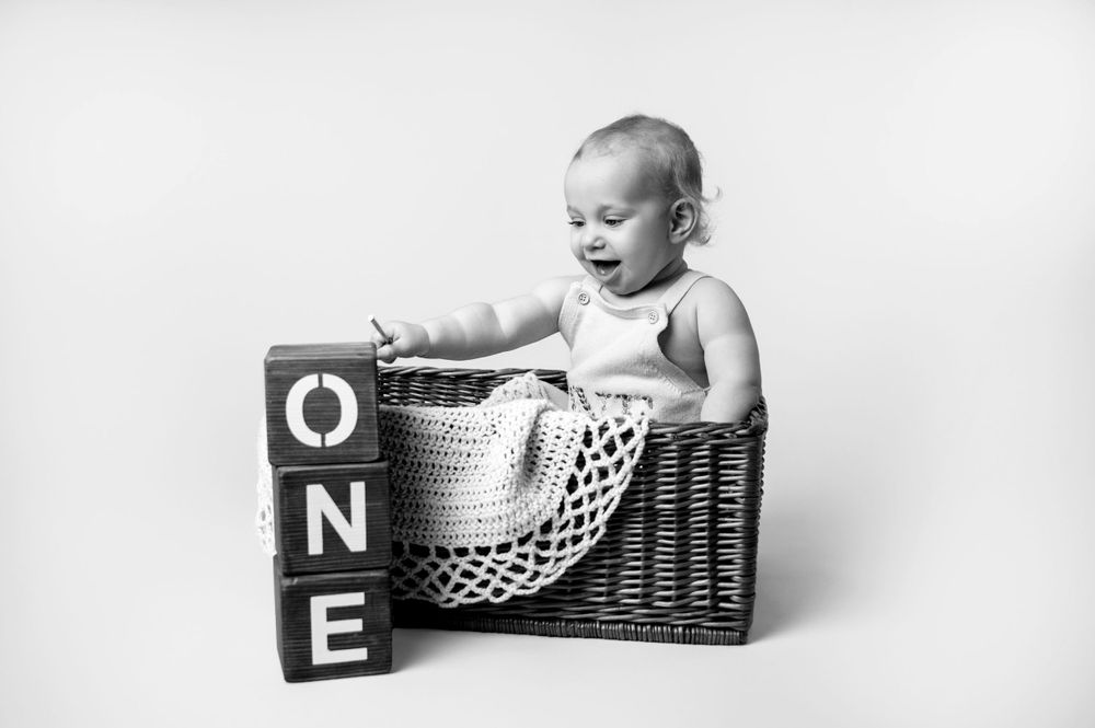 Black and white portrait of baby sitting in wicker basket next to wooden blocks spelling ONE.