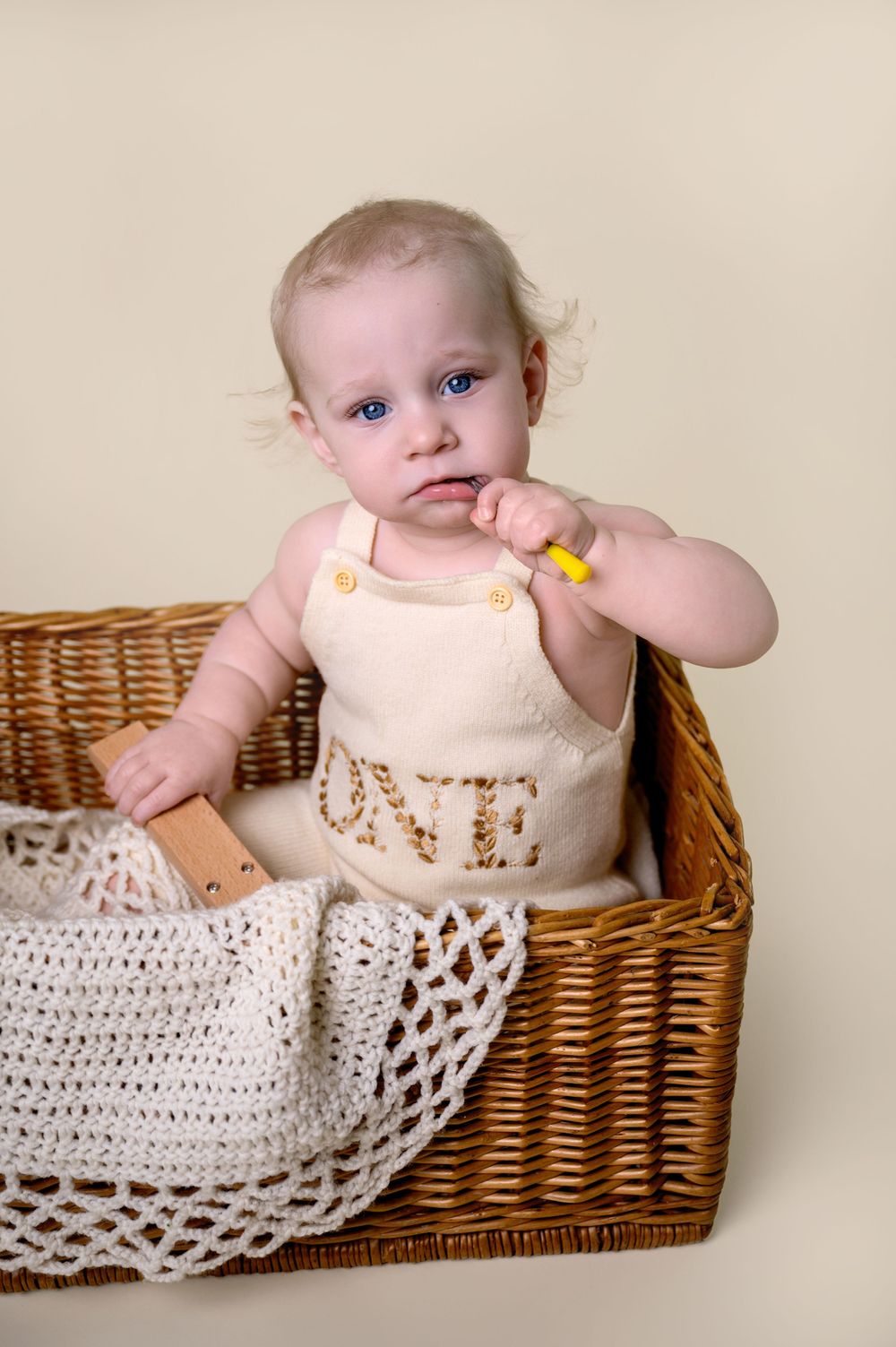 Sweet baby in white outfit sitting in wicker basket with crocheted blanket against beige background.
