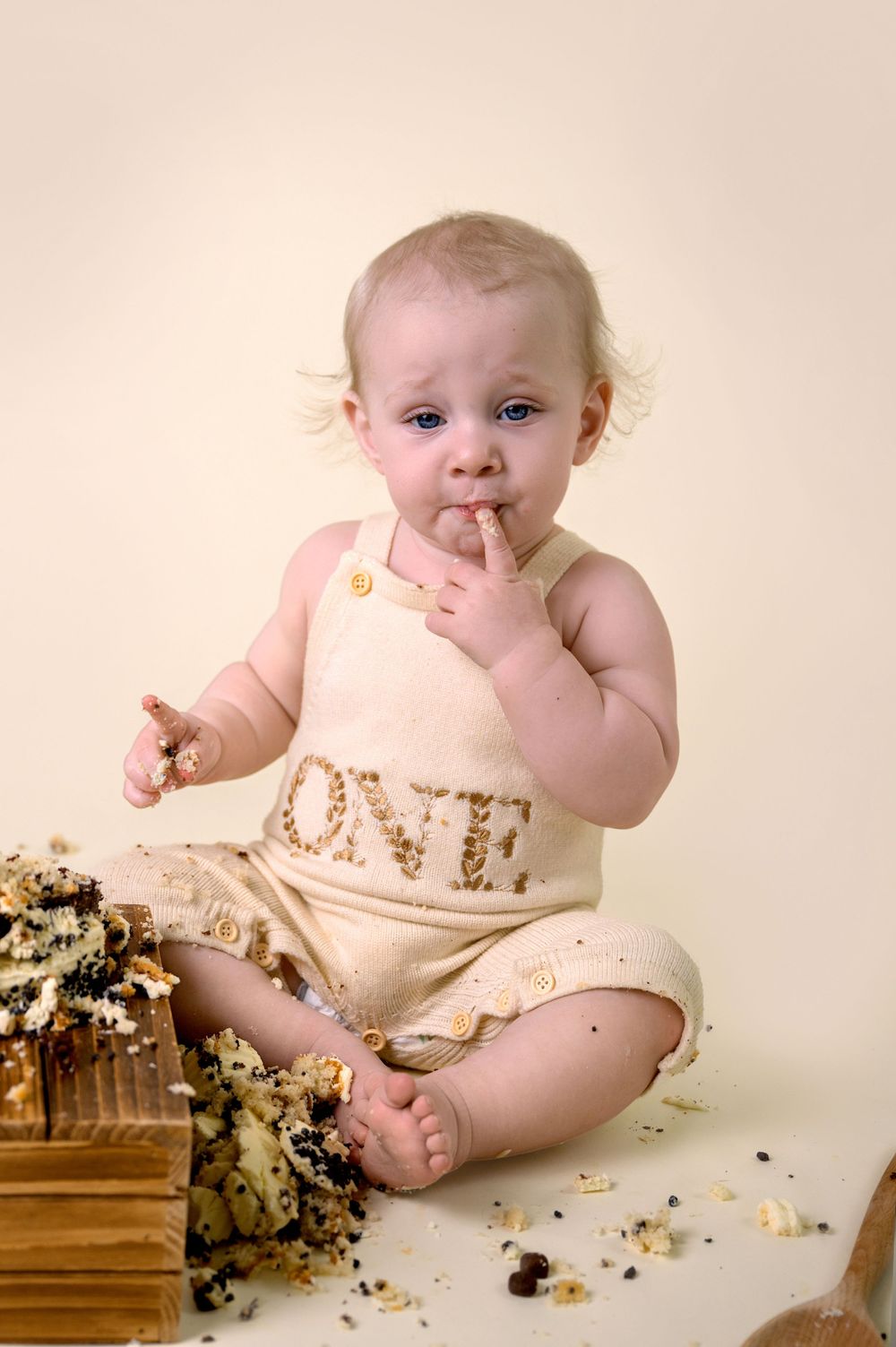 Baby in peach outfit sits on wooden surface with crumbled cake during first birthday cake smash photoshoot.