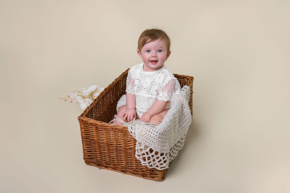 Baby wearing white lace dress sitting happily in wicker basket against neutral background.