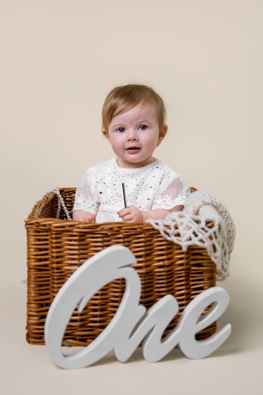 Baby in white dress sitting in wicker basket with ONE sign for first birthday photos.