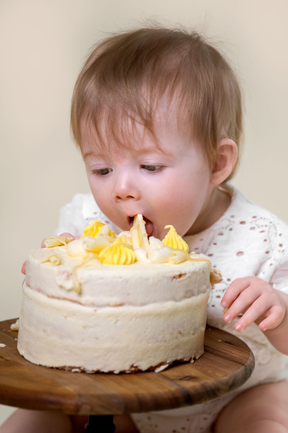Baby leaning towards white birthday cake with yellow frosting on wooden board.
