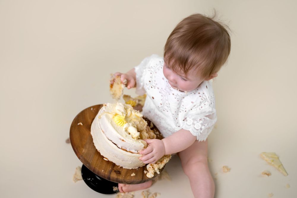 Baby in white outfit sits on floor messily demolishing and playing with a smash cake during first birthday celebration.