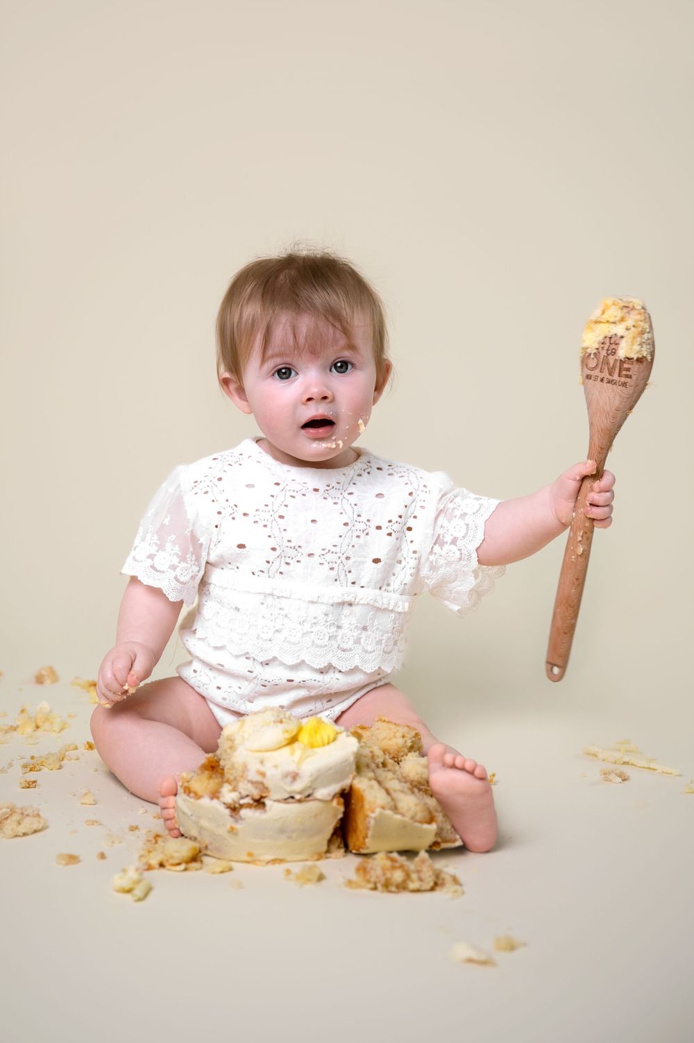 A small child in a white outfit holds a wooden spoon and sits next to a messy cake on the floor.