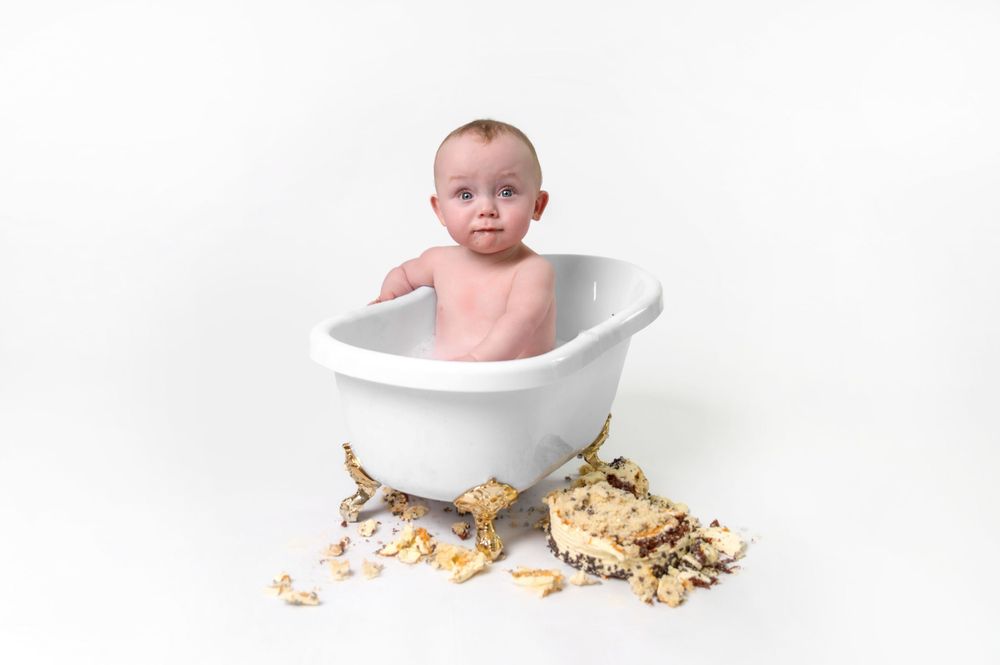 Small baby sitting in white bathtub surrounded by scattered chocolate chip cookies on white background.