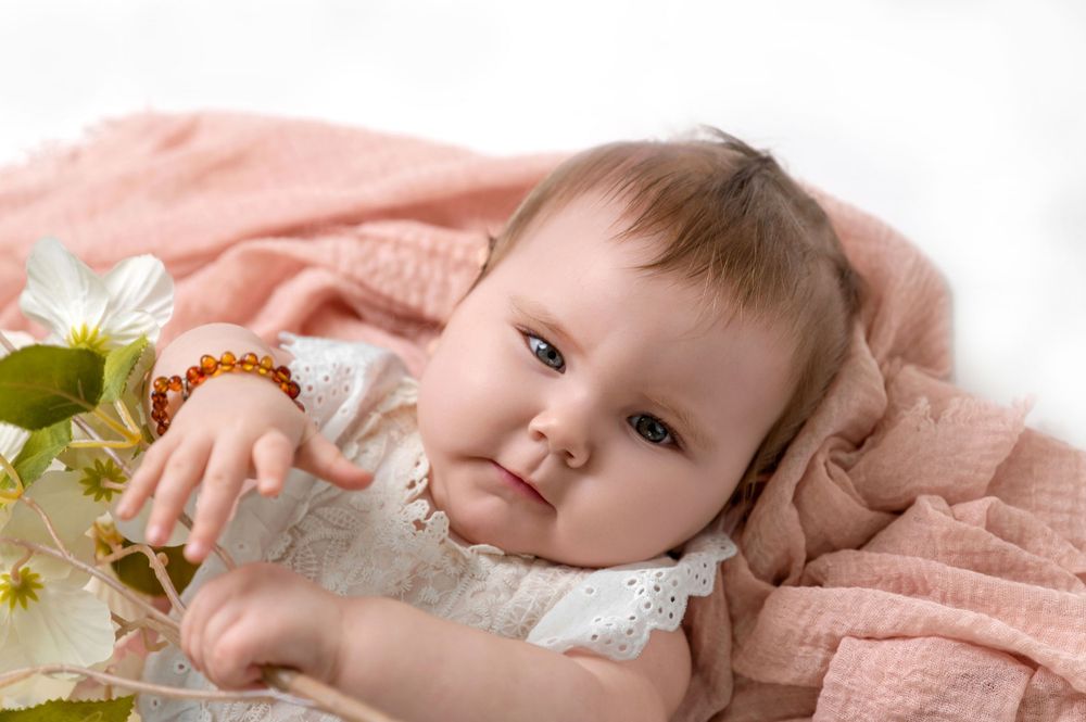 A baby in lace dress lays on soft pink blanket with white flowers, gazing at camera with sweet expression.
