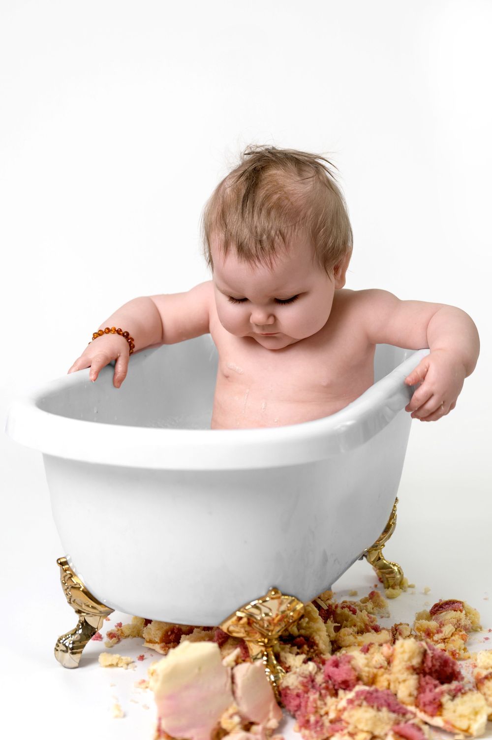 A baby sits in a white bathtub with crumbled cake and frosting scattered below on a white background.