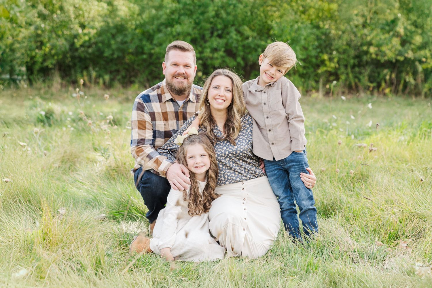 A family kneels together in a sun-lit meadow wearing coordinated casual outfits during an outdoor photo session.