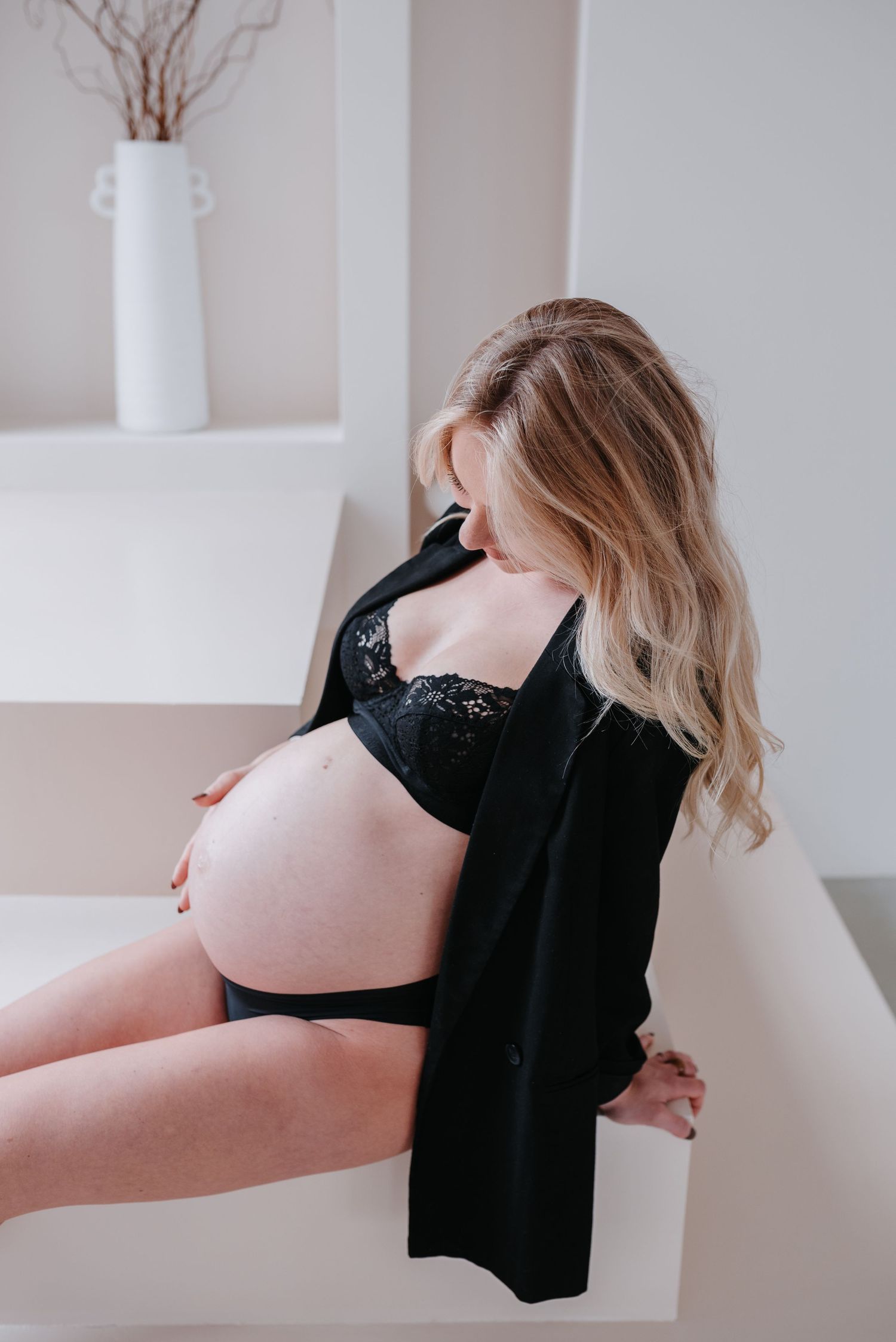 A pregnancy photo of someone in black lingerie sitting on a white surface with a decorative vase in background.