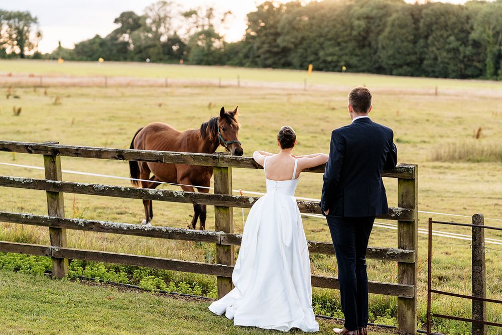 A couple in wedding attire stands together at a rustic wooden fence overlooking a pasture with horses.