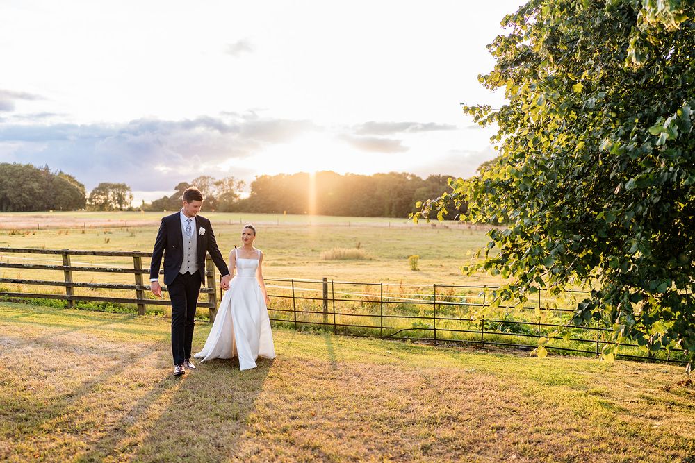 A romantic couple walks along a rural path during golden hour with sun flare and rustic fencing in the background.
