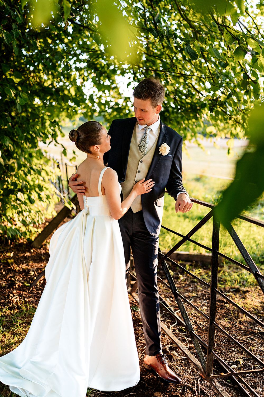 A couple in wedding attire embrace on a rustic path under green foliage during a golden sunset.