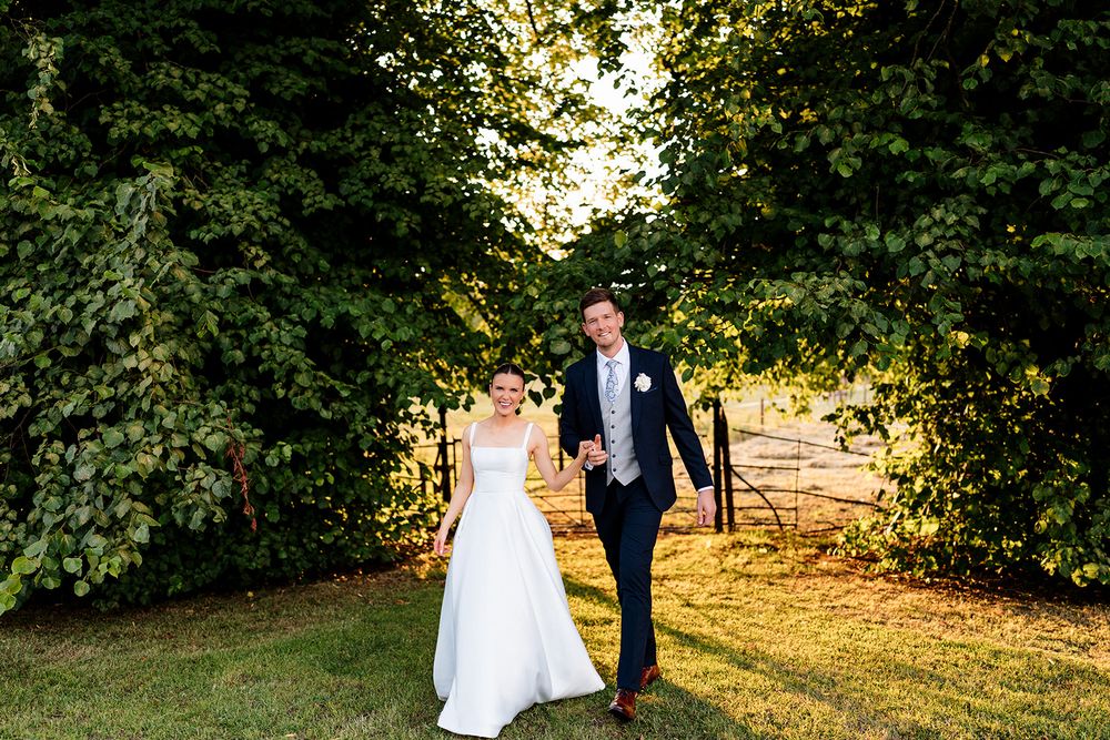 A couple in wedding attire walks through a natural garden arch during golden hour sunset light.
