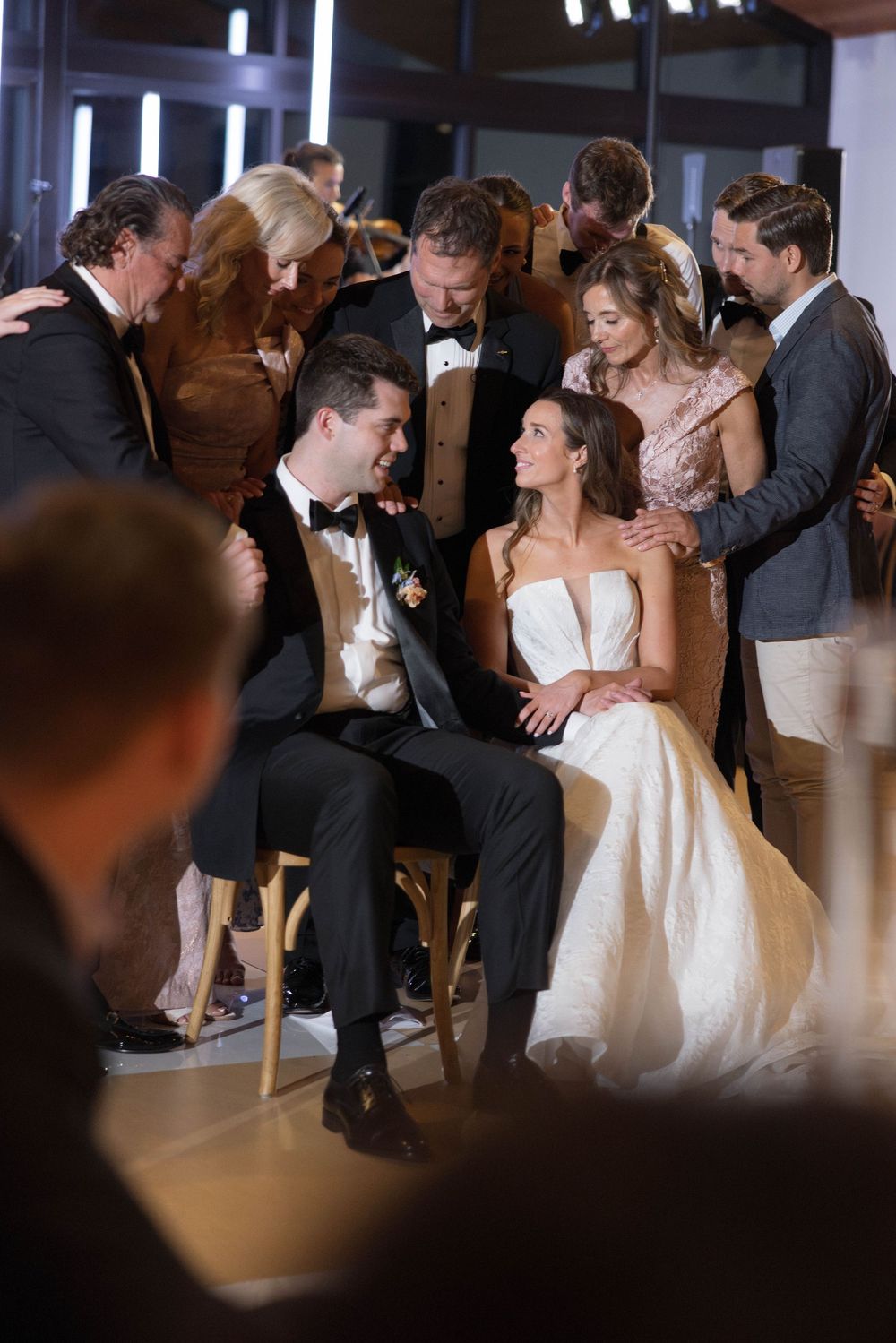 Wedding couple surrounded by guests share an intimate moment during reception while seated on chairs.