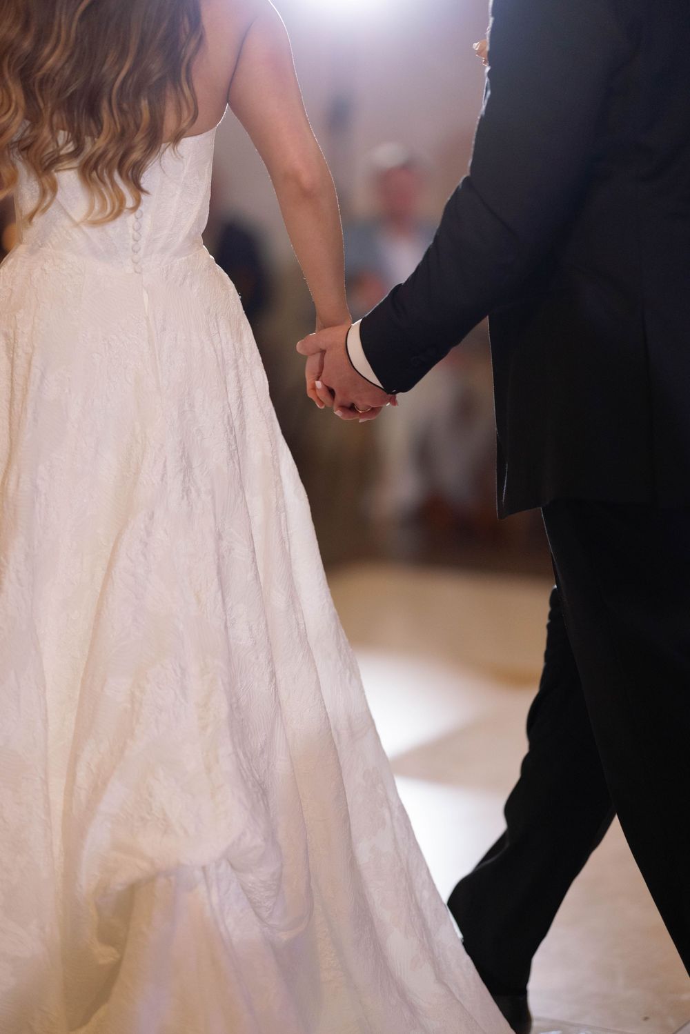 Close-up of bride in white lace gown holding hands with groom in black suit during wedding ceremony.