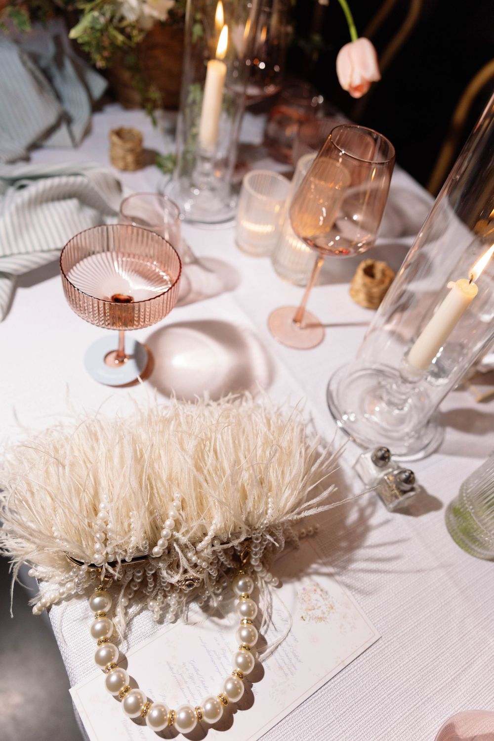 Elegant table setting with pink glassware, white candles, and pearl necklace on white linen tablecloth.