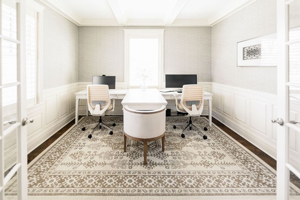 A modern home office space featuring two desks, ergonomic chairs, and an ornate area rug on hardwood floors.