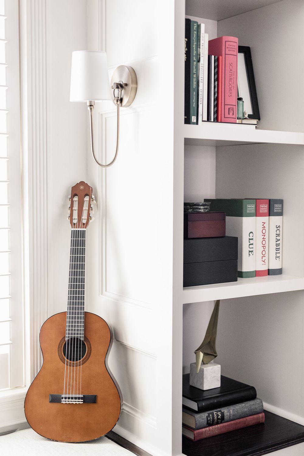 Classical guitar leans against wall beside built-in bookshelf with books and wall sconce light fixture.