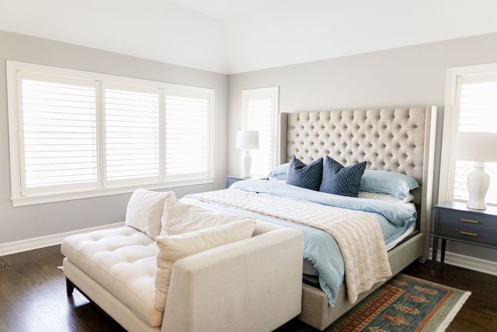 Bright modern bedroom with neutral tones featuring a tufted headboard, blue accents, and natural light from windows.