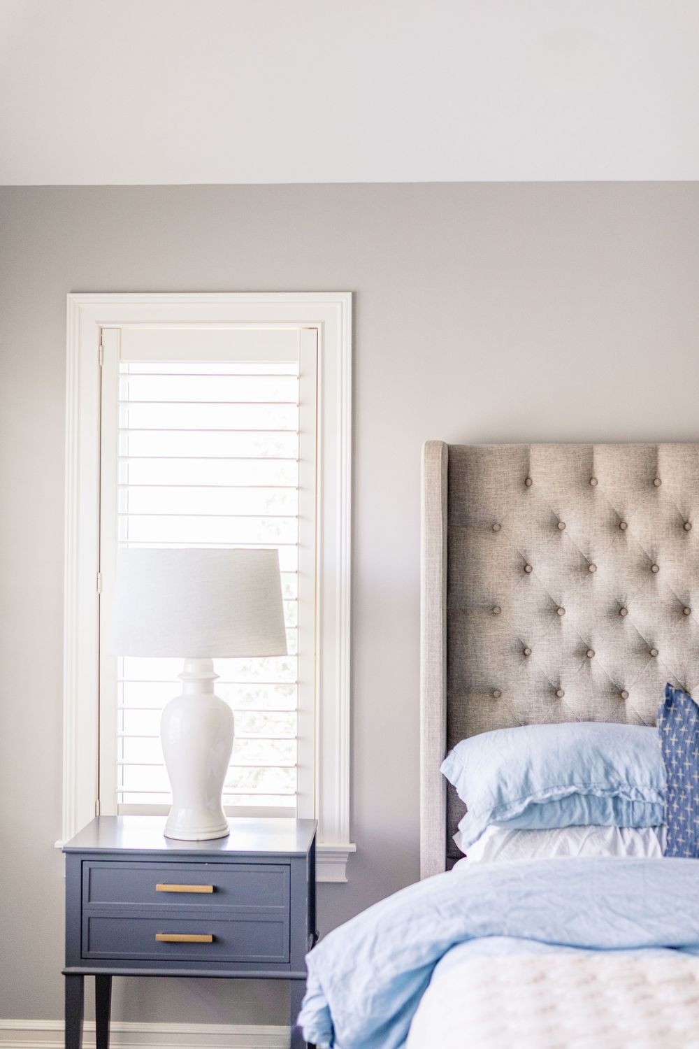 A serene bedroom with a gray tufted headboard, blue bedding, and a white lamp on a navy nightstand beside a window.