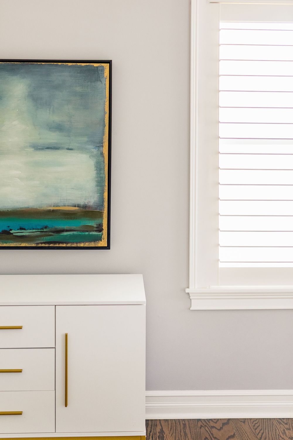 Modern white dresser with brass handles beneath an abstract landscape painting in blue and gray tones.