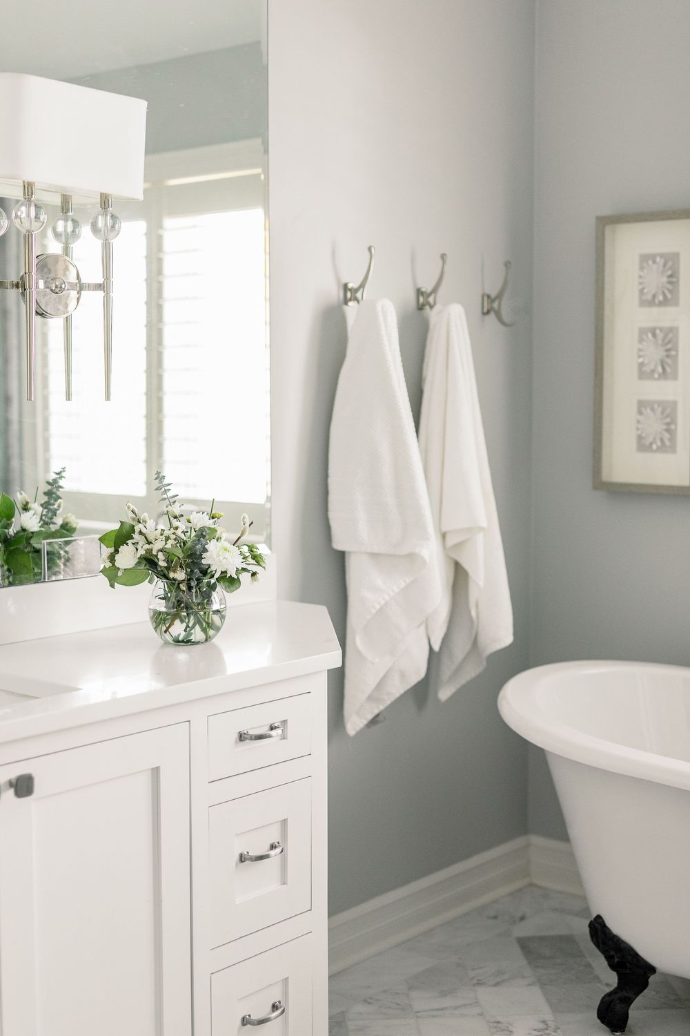 Elegant white bathroom with marble floors, freestanding tub, white vanity, and fresh flowers in a bright window setting.