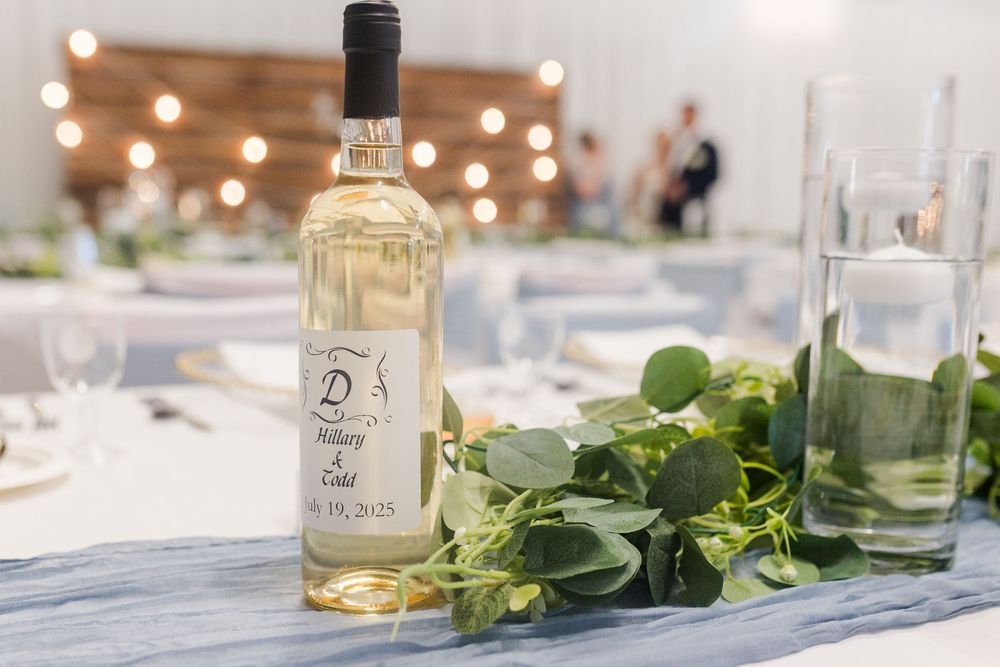 A white wine bottle sits on an elegantly set table with fresh herbs and glasses under warm string lights.