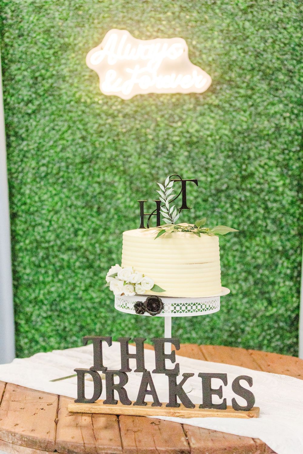 A simple white wedding cake on a wooden stand with a green backdrop and 'THE DRAKES' text displayed below.