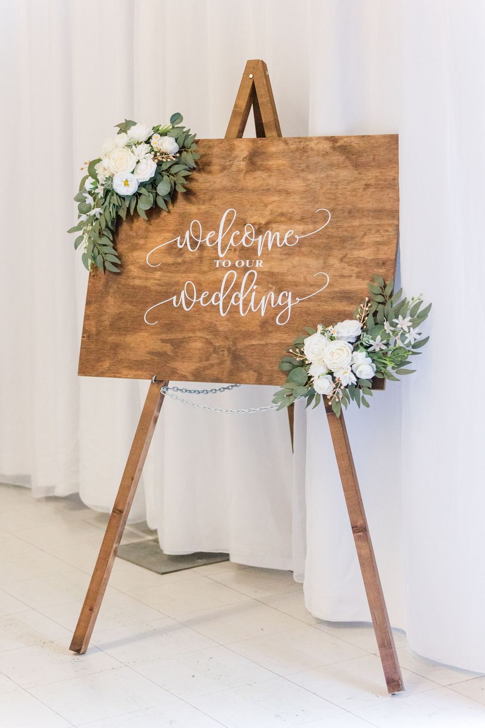 Rustic wooden welcome sign decorated with white flowers and greenery against a light backdrop.