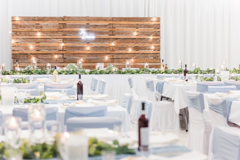 Elegant wedding reception setup with white chairs, floral greenery, and a rustic wooden backdrop with string lights.