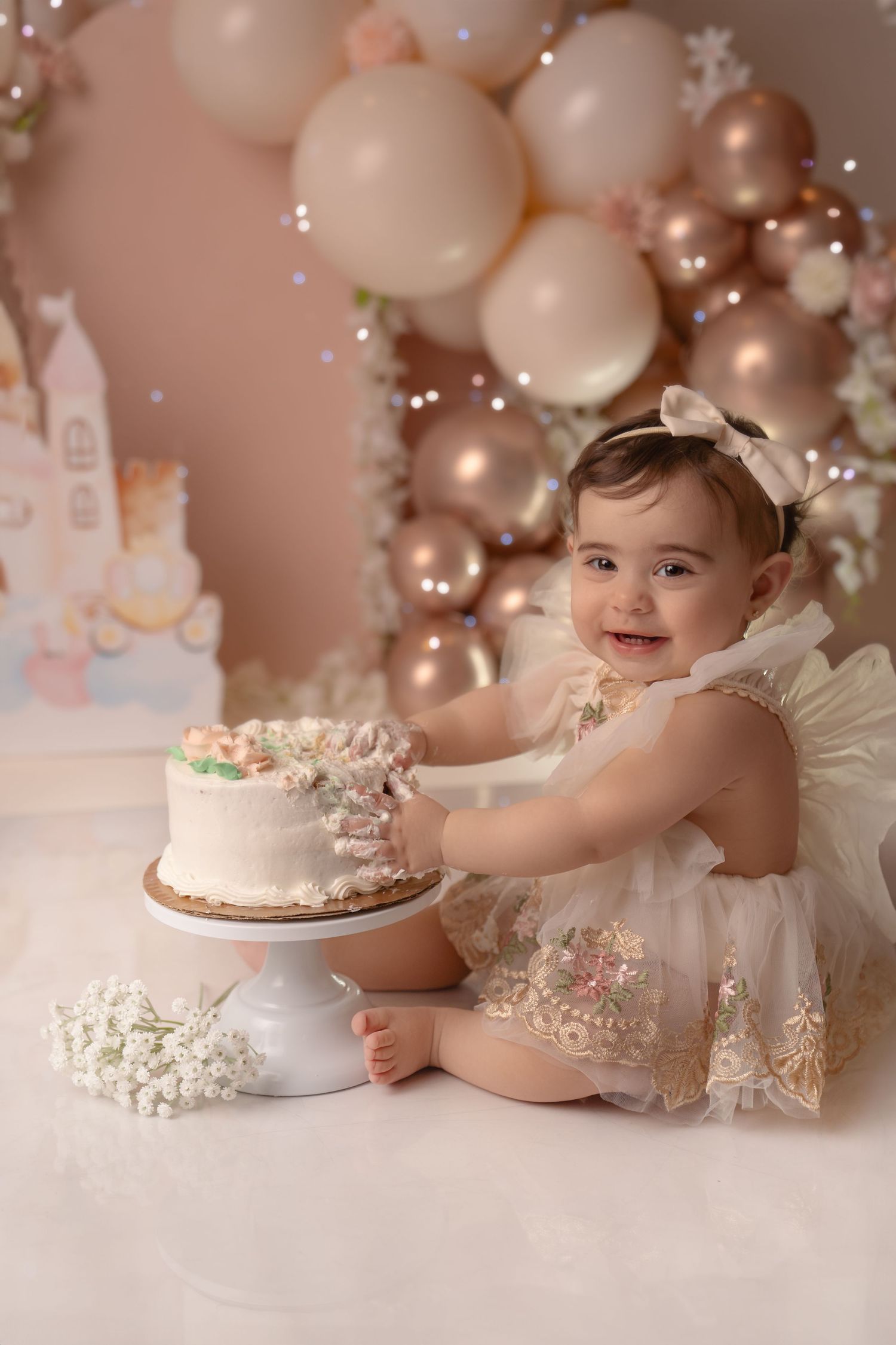 Baby in white dress reaching for small white birthday cake with pink and gold balloon decorations in background.