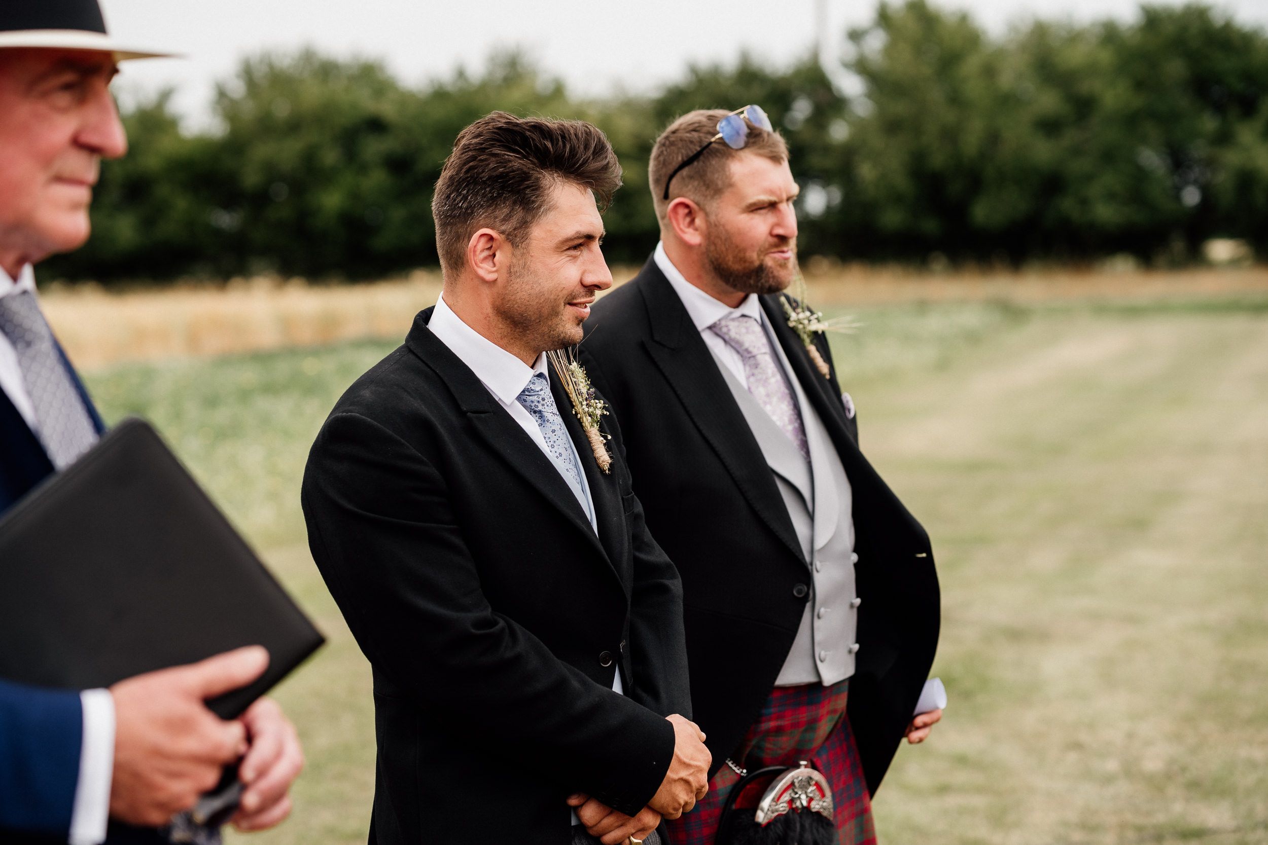 Wedding guests sit outdoors on wooden chairs during a summer ceremony in a grassy field.