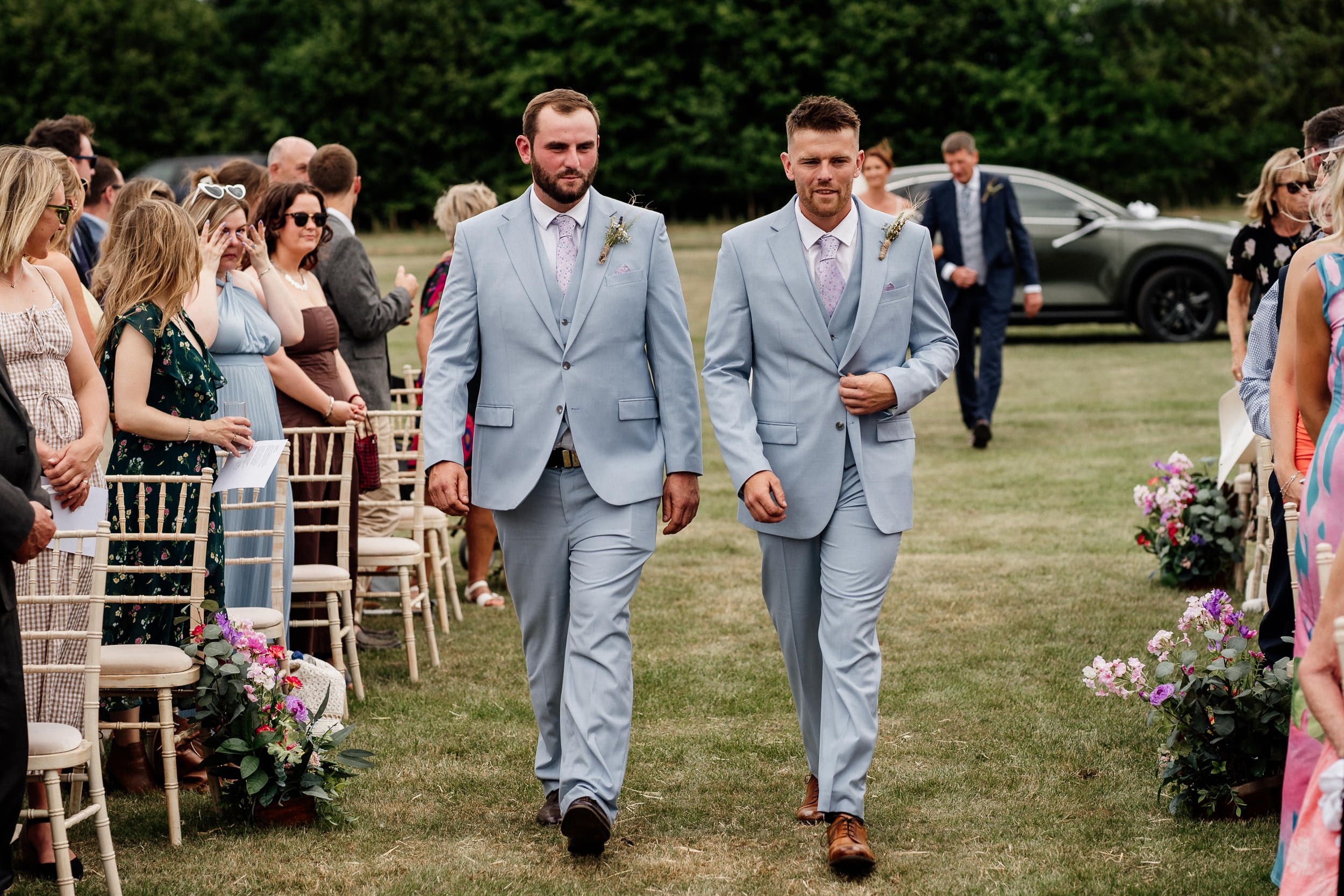 A wedding party in light blue suits and pink dresses walks through a grassy outdoor venue during a summer celebration.