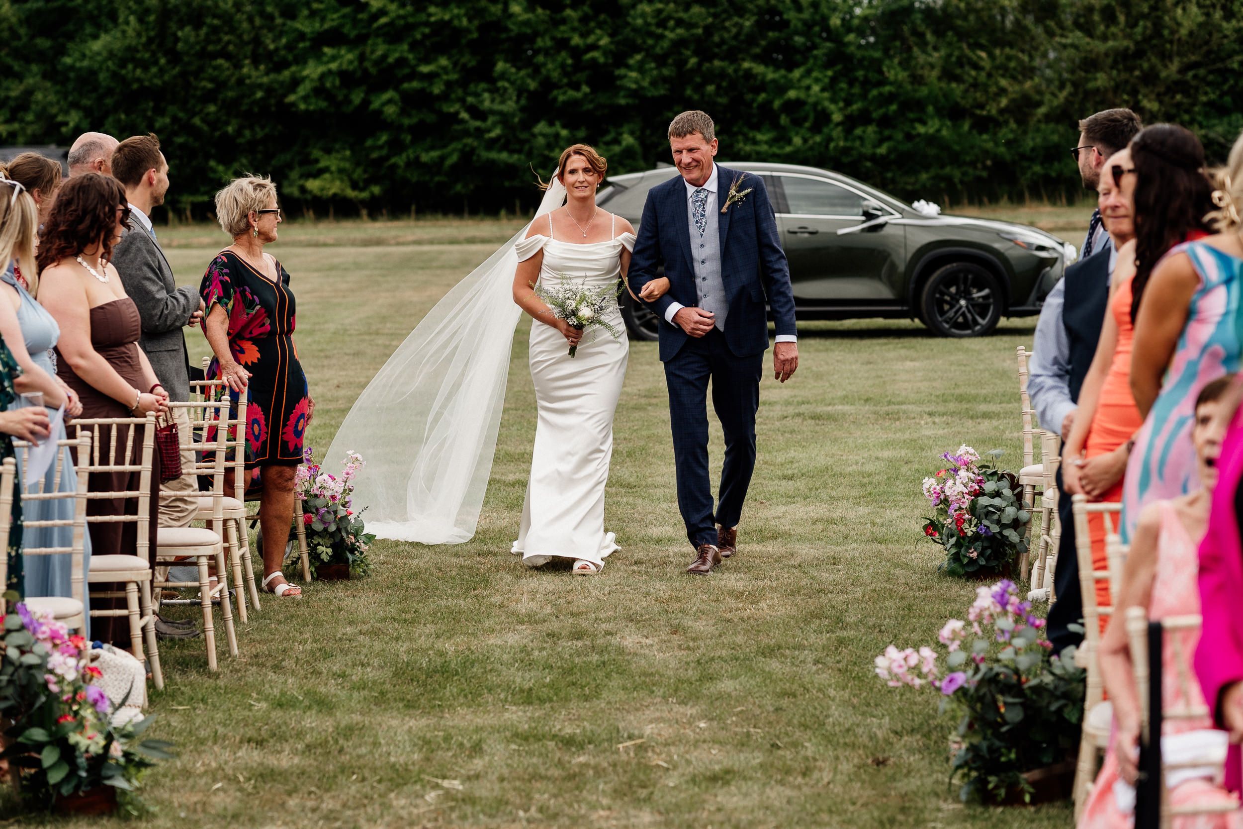 A group gathers outdoors for a wedding ceremony with white chairs arranged in rows and floral decorations on a summer day.