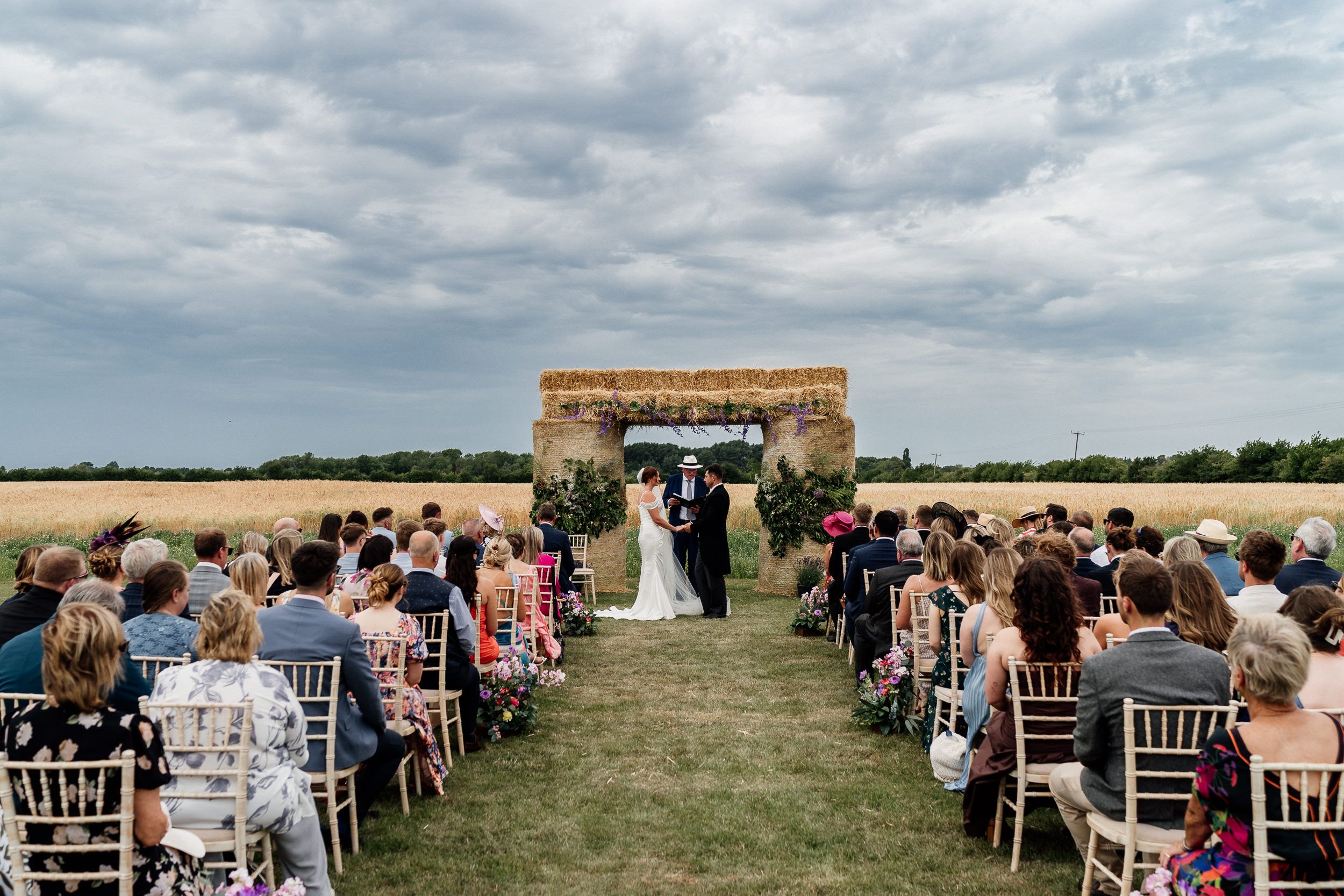 An outdoor wedding ceremony with guests seated in rows of chairs on a green grassy field under cloudy skies.