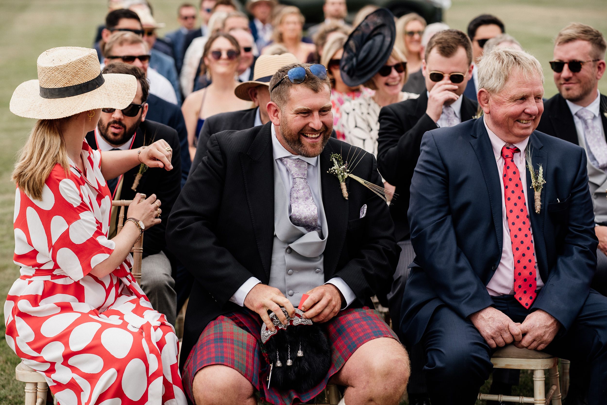 Guests share a laugh while seated in rows during an outdoor event wearing business attire and sunglasses.