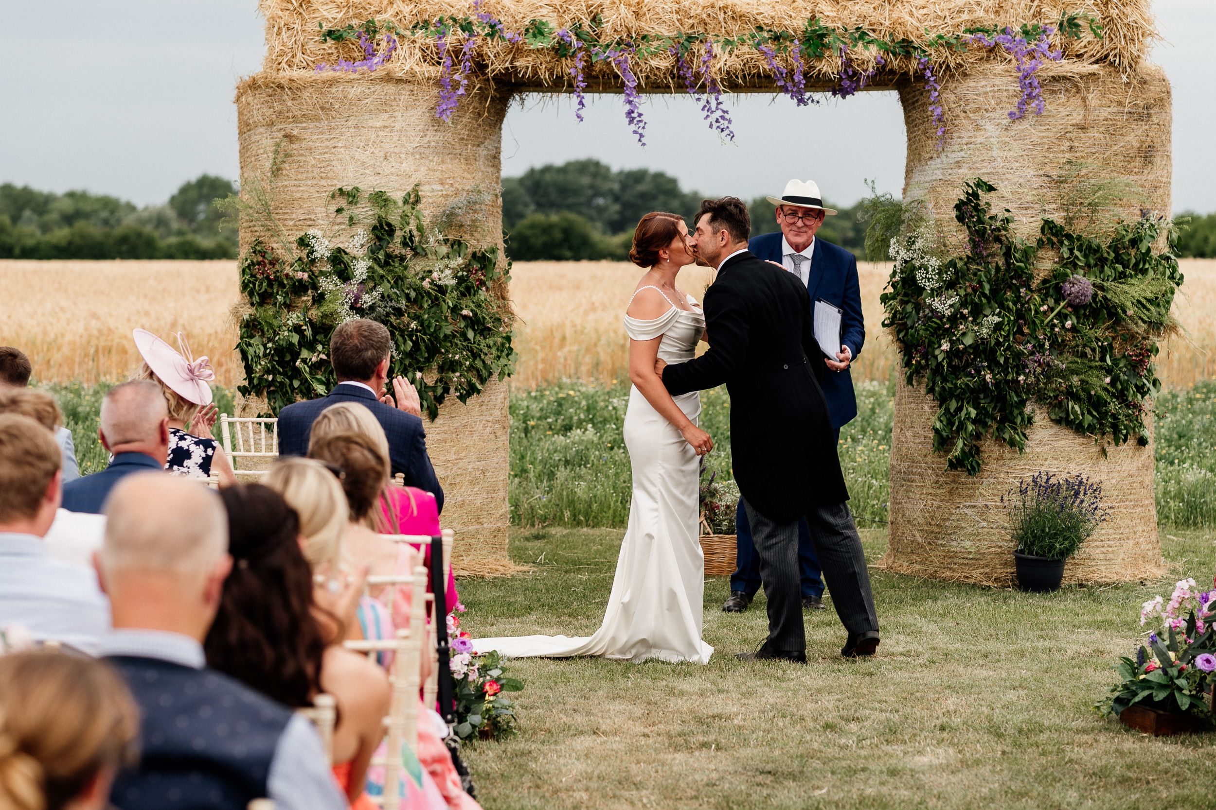 Outdoor wedding ceremony takes place under rustic stone archway decorated with greenery in countryside setting.