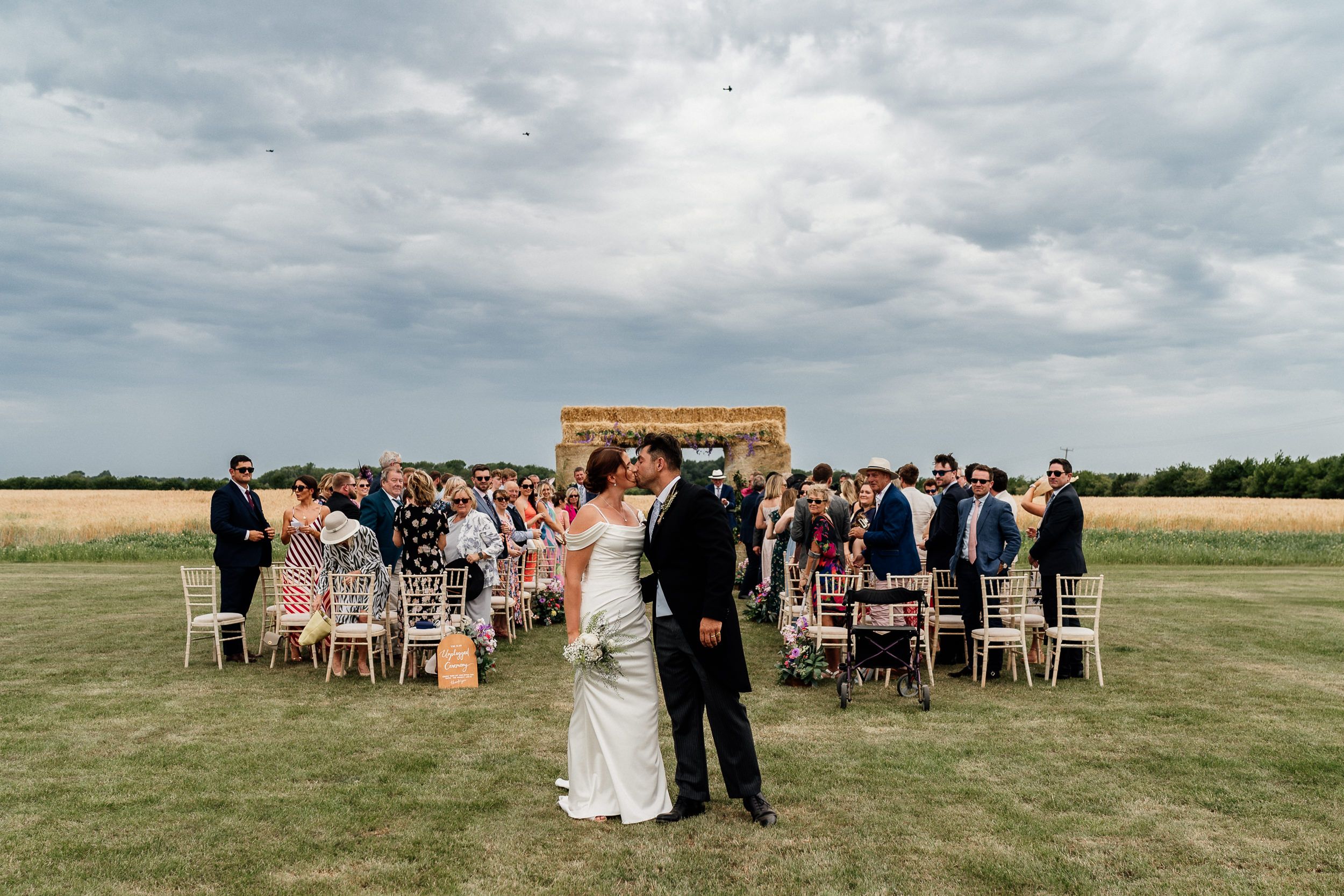 Wedding ceremony guests gather on a scenic grassy field under cloudy skies as chairs line up for the outdoor celebration.