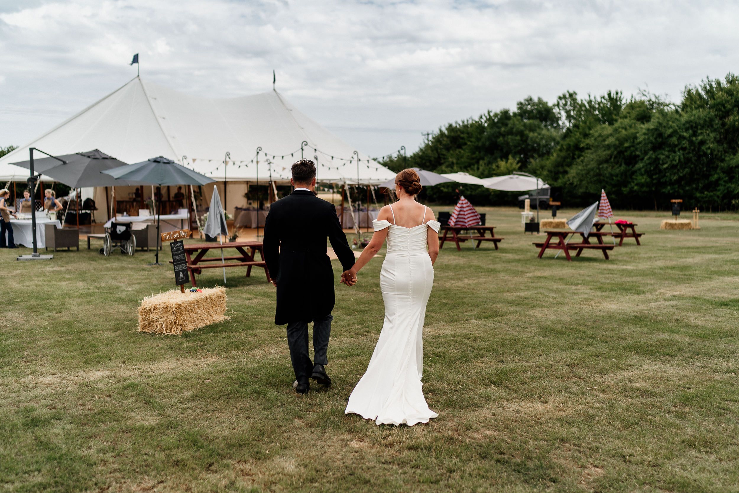 A couple walks hand in hand towards an outdoor wedding tent setup with picnic tables on a grassy field.