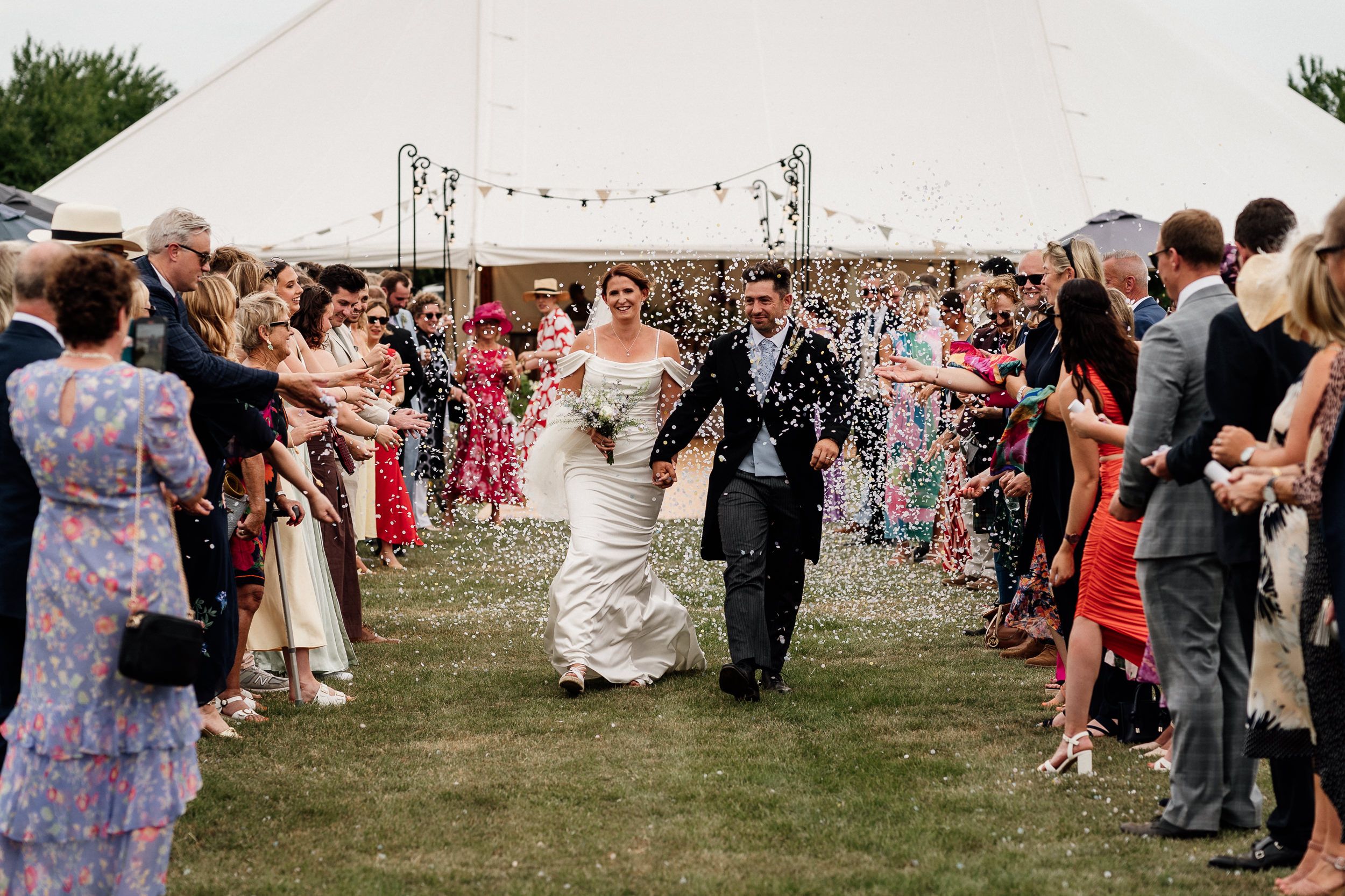 Wedding couple walks through confetti shower thrown by guests outside white tent at outdoor celebration.